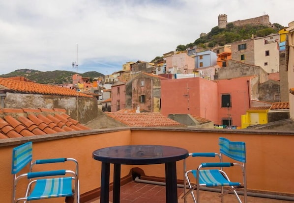 A terrace with two blue chairs and a round table, overlooking a colorful hillside town with historic buildings and a castle on a hill. - View of the "Sa Costa" Castle (Bosa)