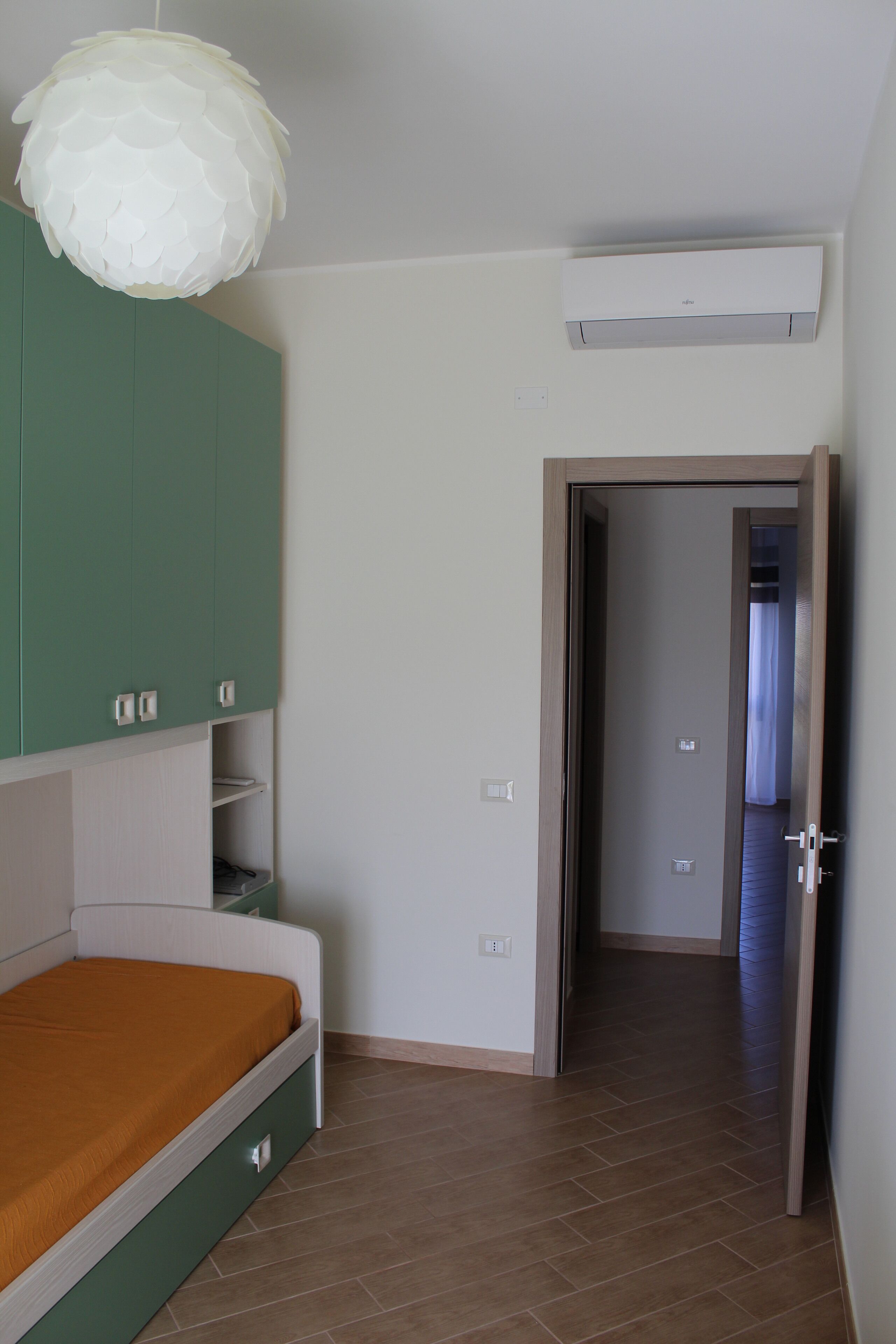 A bedroom with a bed, a green wardrobe, a white air conditioner, and a door leading to another room.