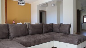 Un salon moderne avec un canapé d’angle, un luminaire suspendu et une vue sur une autre pièce.
