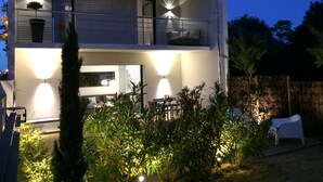 A modern two-story house with a balcony, outdoor seating, and well-lit garden areas.
