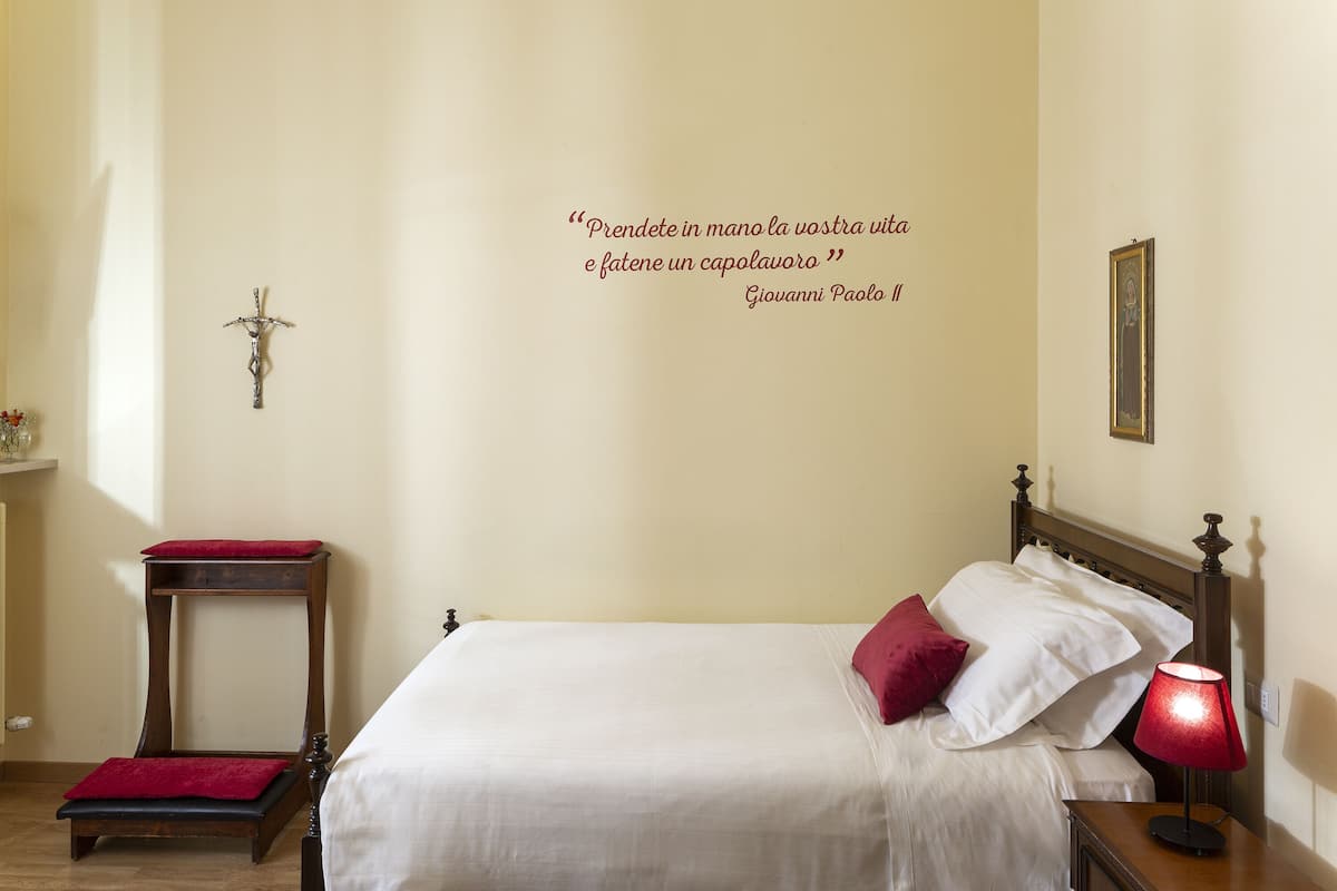 A bedroom with a bed, a bedside table, a nightstand, a wall-mounted cross, a framed picture, and a quote on the wall.