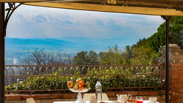 A table with a fruit bowl, teapot, cups, and pastries, set against a scenic mountain backdrop. - Hotel Sacro Cuore (Perugia)