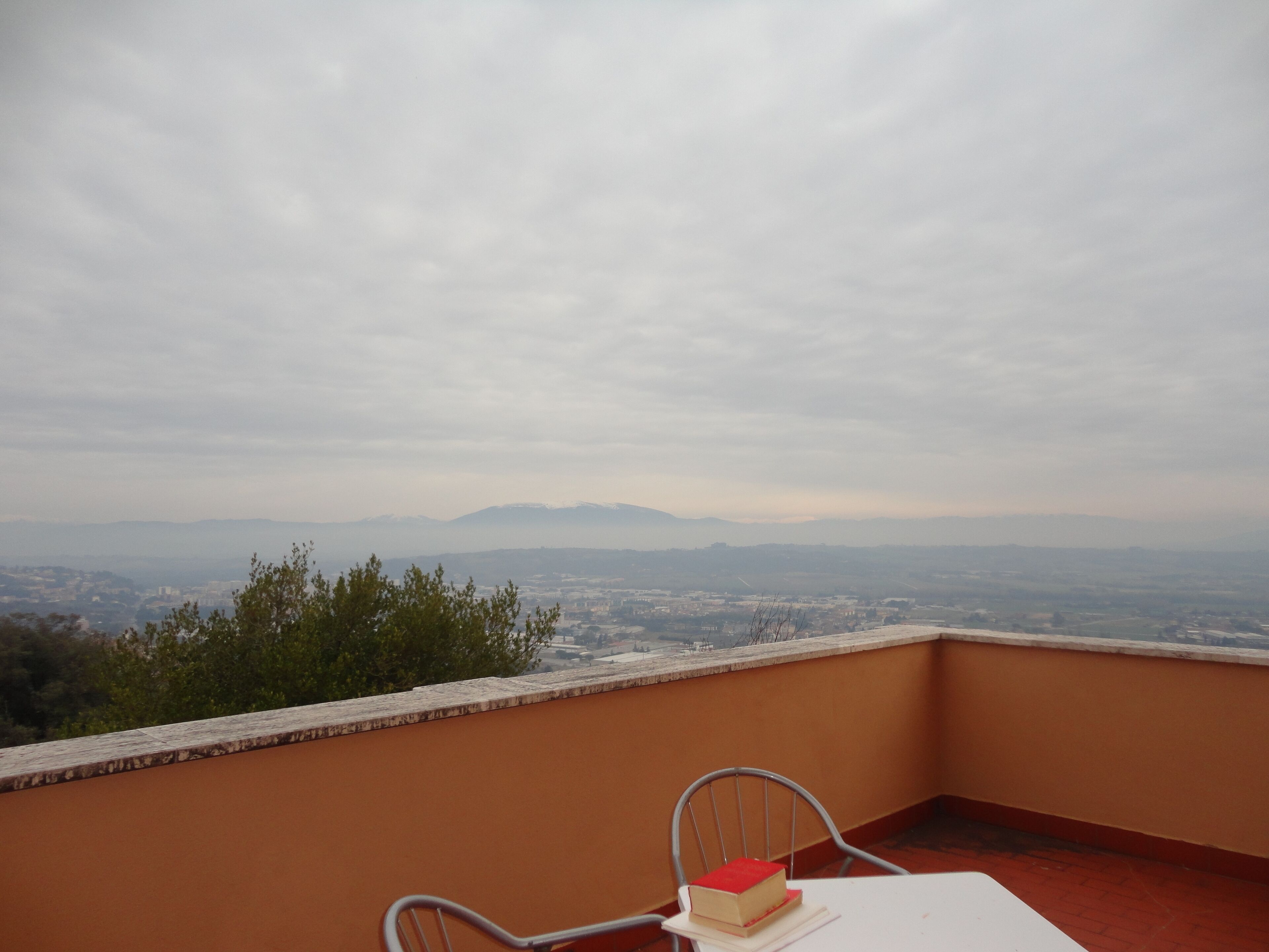 Un balcone con tavolo e sedia, affaccia su un panorama urbano con montagne in lontananza.