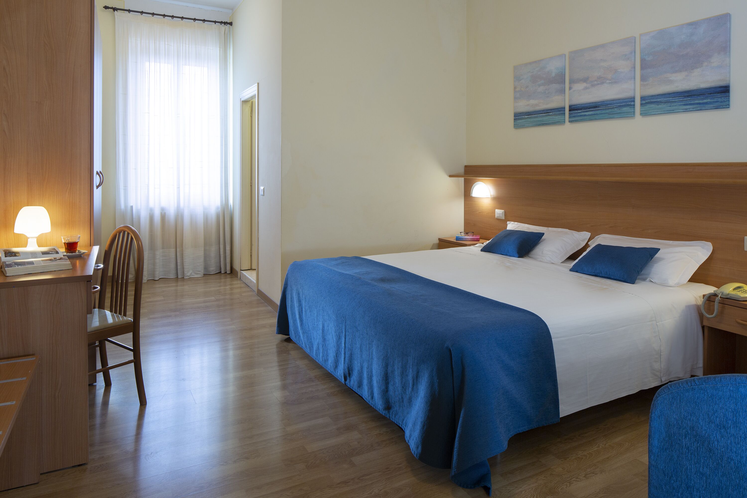 Ein Hotelzimmer mit einem großen Bett, einem Schreibtisch mit Lampe, einem Sessel und Blick auf das Meer durch das Fenster.