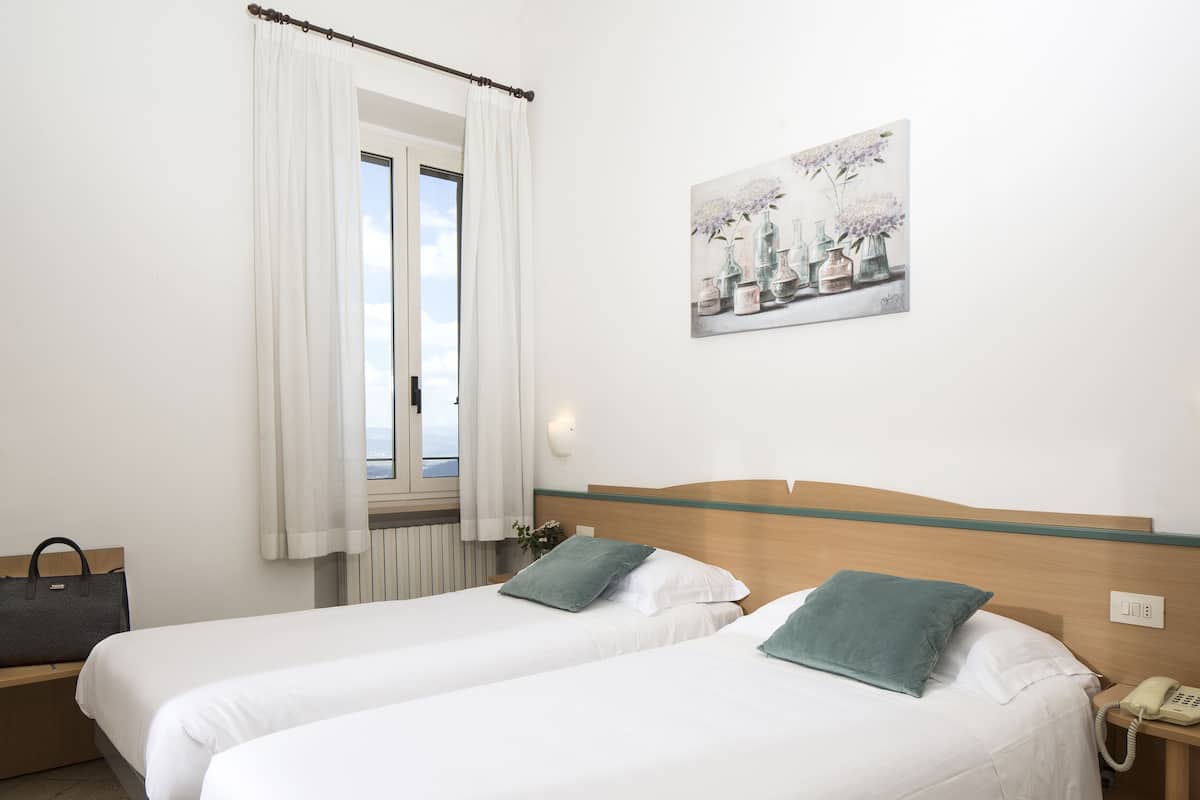 A hotel room with two single beds, a wooden headboard, a bedside table with a telephone, a window with white curtains, and a wall-mounted painting.