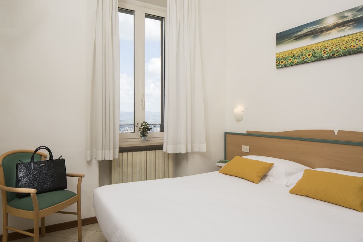 A hotel room with a bed, a chair, a window with curtains, and a painting on the wall.