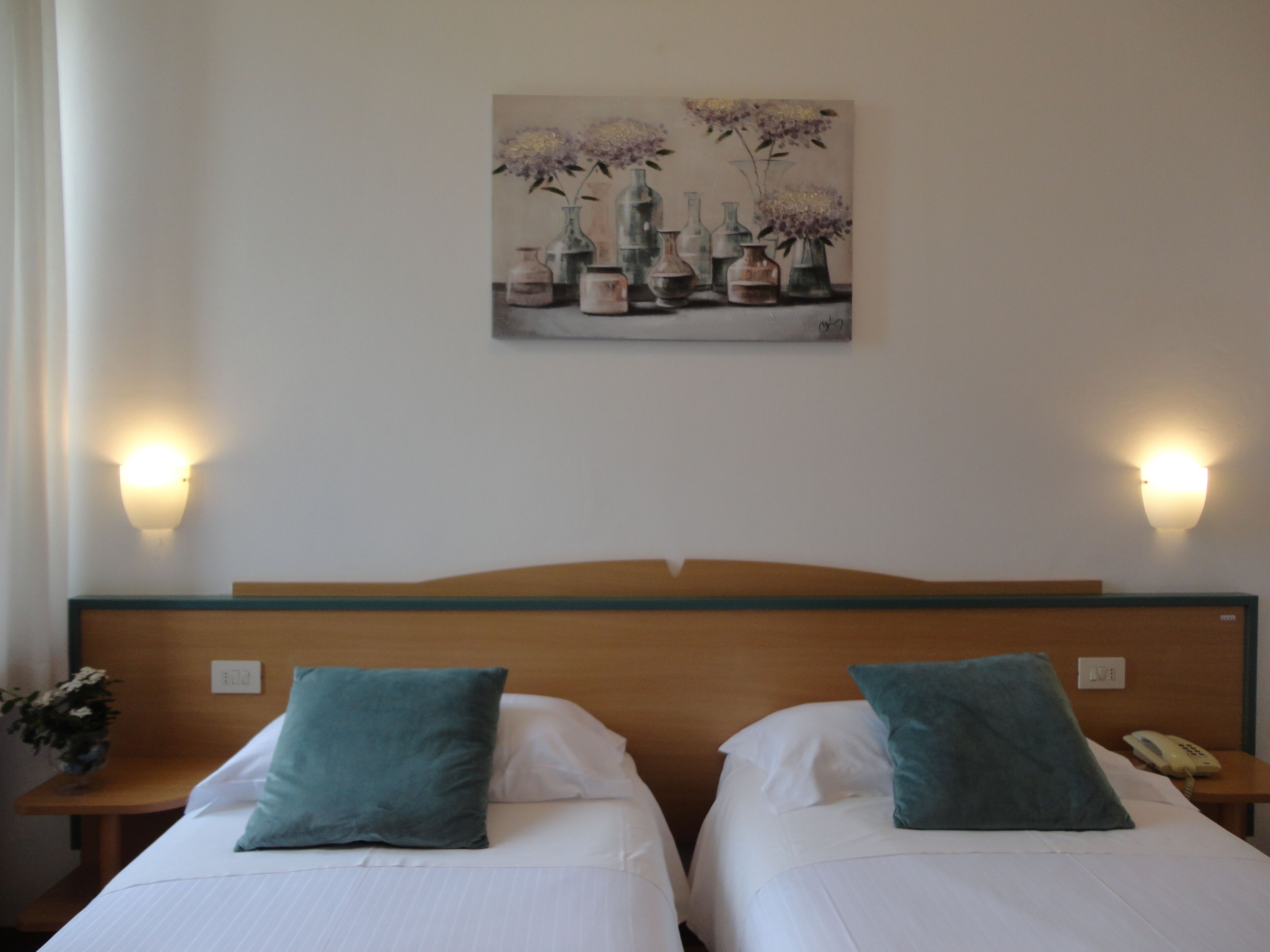 Quarto de hotel com duas camas, cada uma com travesseiro e mesa de cabeceira com abajur. Há um quadro na parede acima das camas.