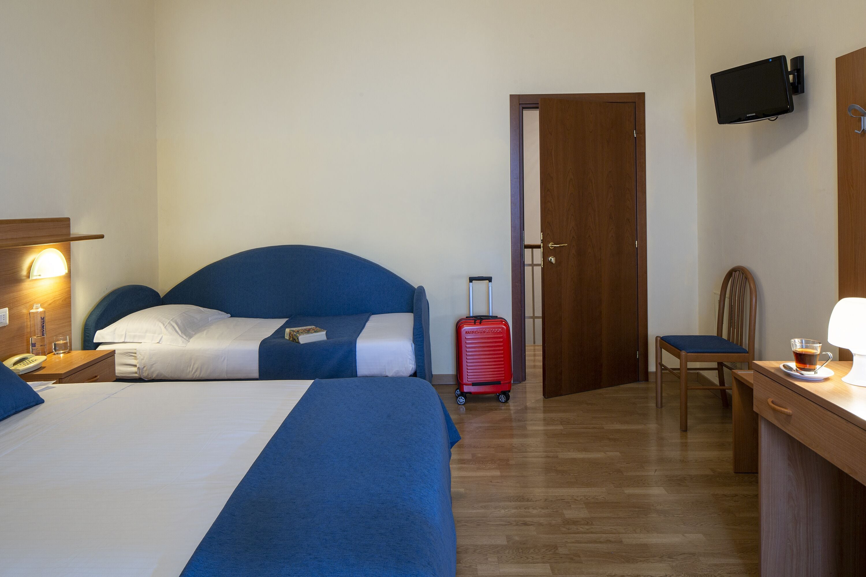 Habitación de hotel con dos camas, un escritorio, una silla, un televisor y una maleta.