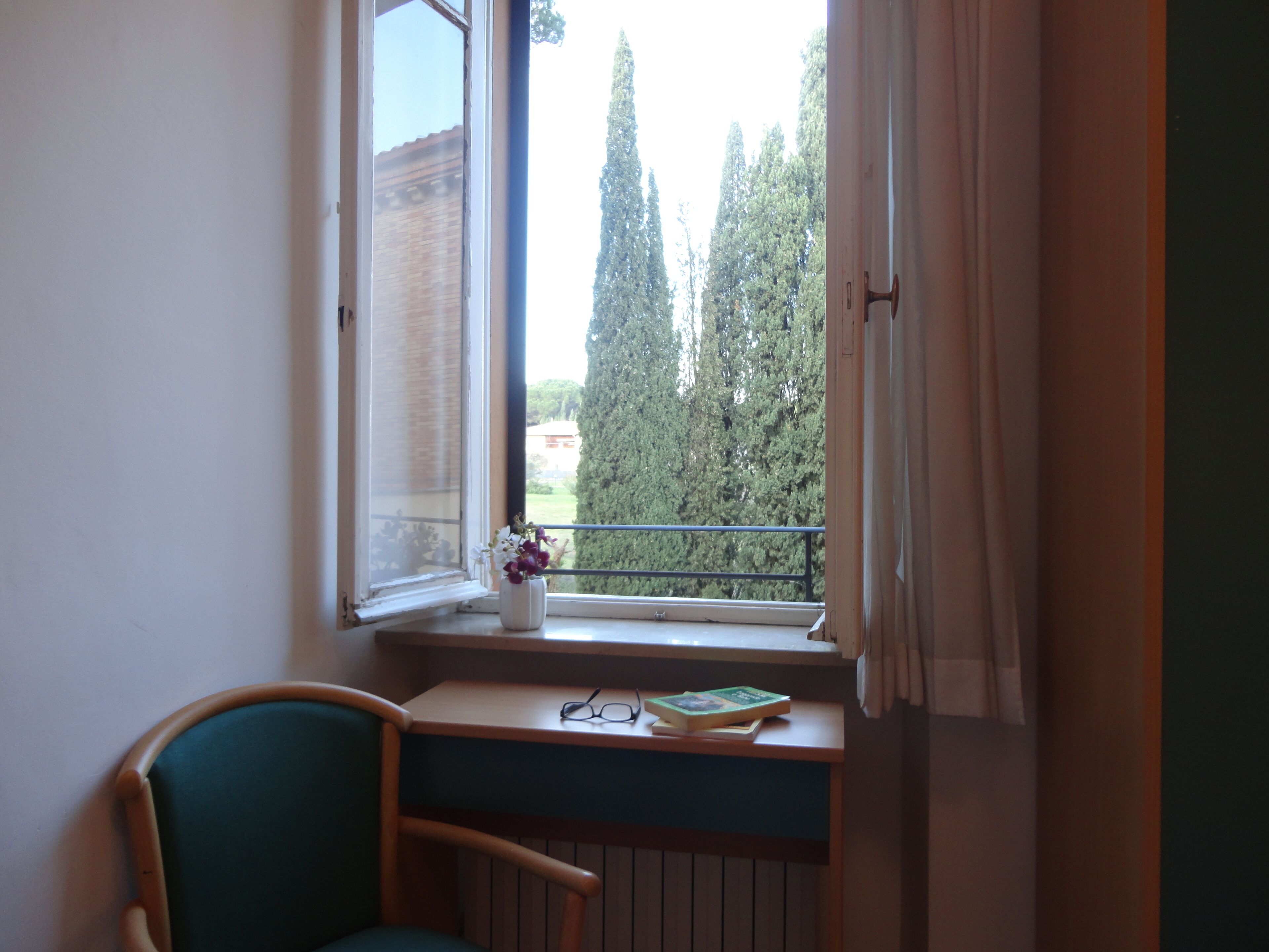 Una stanza con una finestra che si affaccia su un balcone, una sedia e un tavolino con un libro e occhiali.