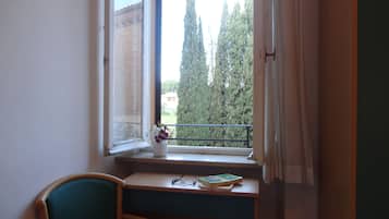 Una stanza con una finestra che si affaccia su un balcone, una sedia e un tavolino con un libro e occhiali.