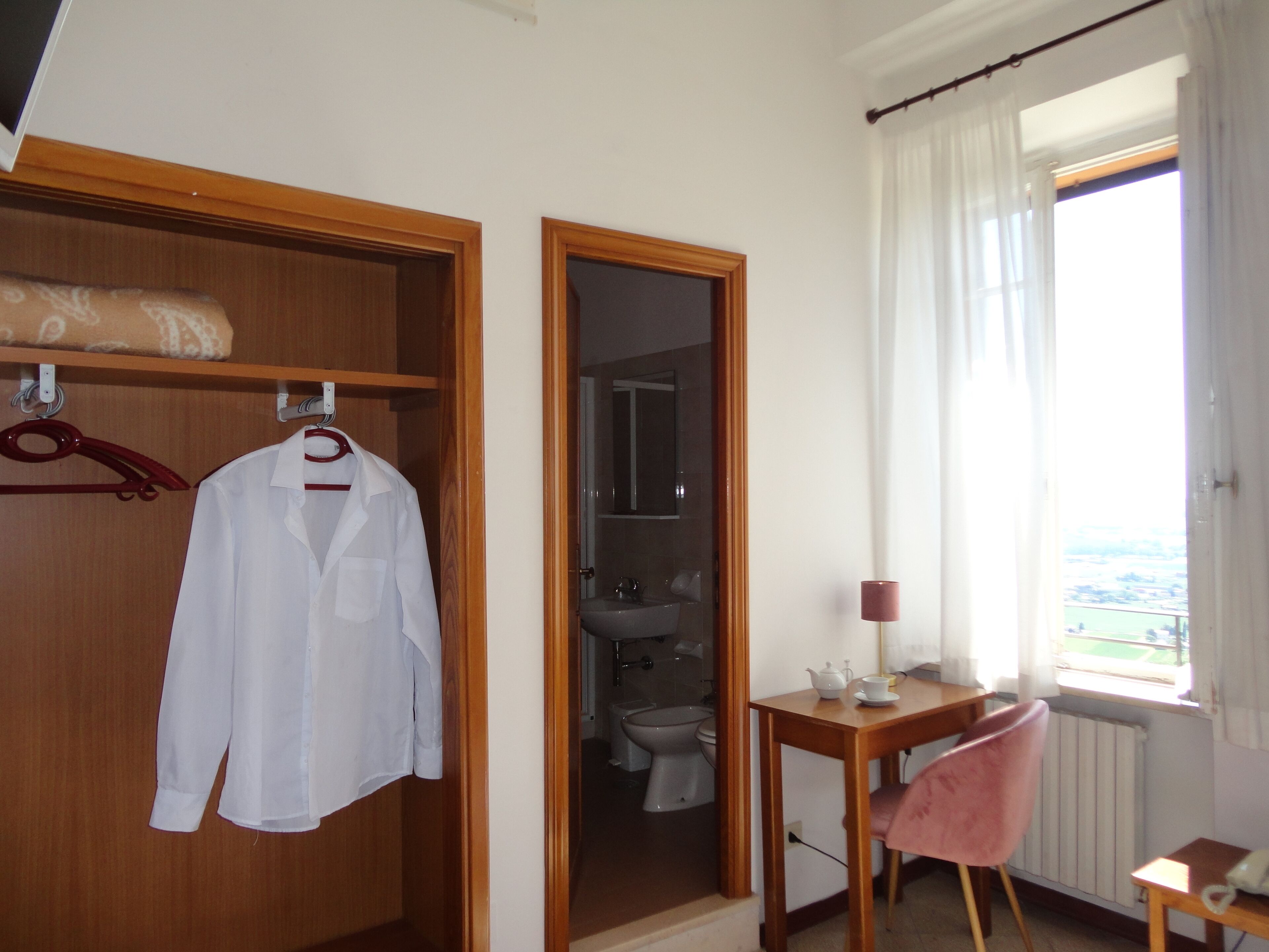 Ein Hotelzimmer mit Holzschrank, einem aufgehängten weißen Hemd, einem Badezimmer mit Toilette und Waschbecken, einem kleinen Tisch mit Lampe und einem Fenster mit Blick.