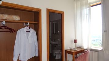 Ein Hotelzimmer mit Holzschrank, einem aufgehängten weißen Hemd, einem Badezimmer mit Toilette und Waschbecken, einem kleinen Tisch mit Lampe und einem Fenster mit Blick.