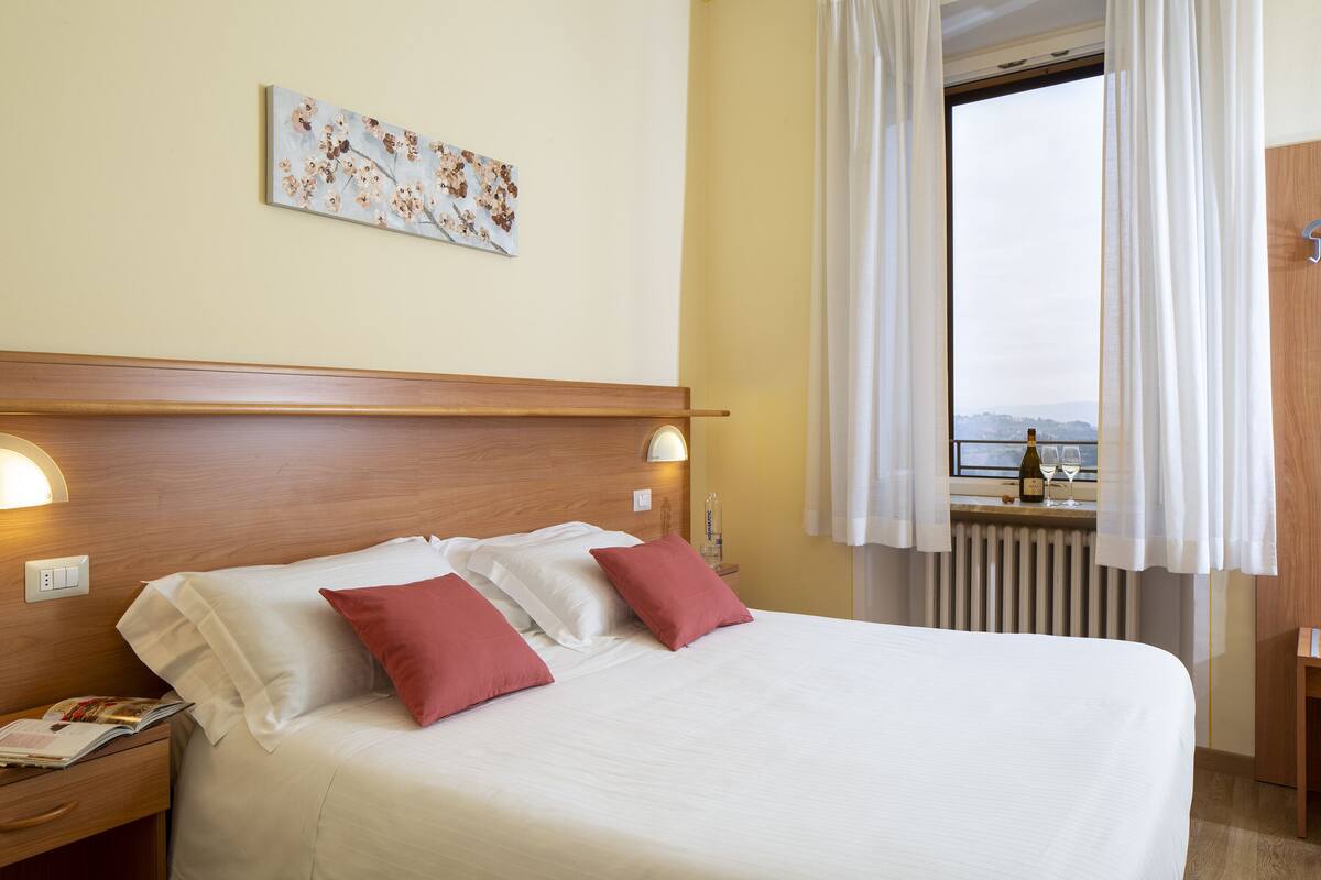 A hotel room with a bed, a nightstand, a window with curtains, and a wall-mounted light fixture.