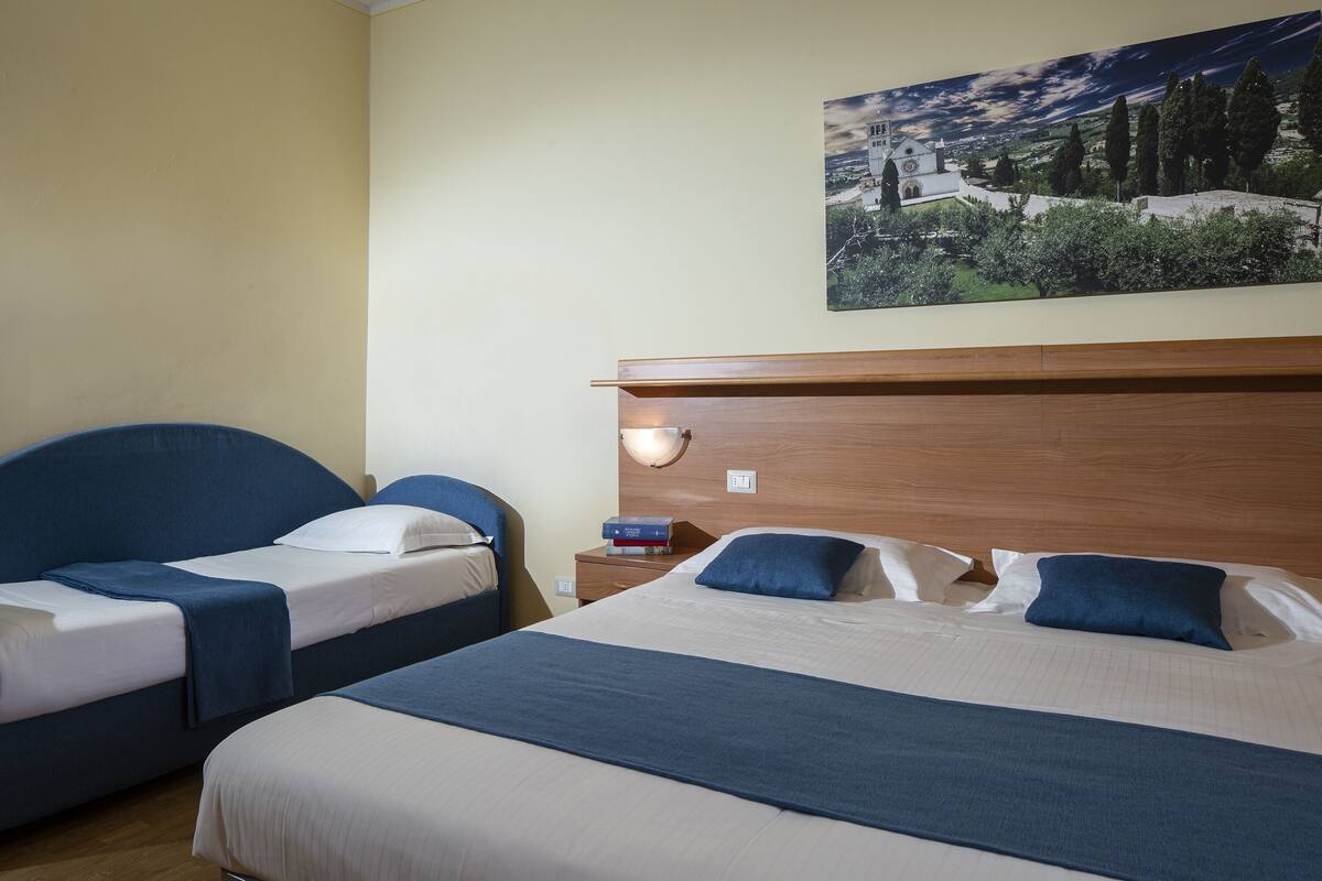 A hotel room with two single beds, a wooden headboard, a nightstand, and a wall-mounted lamp.