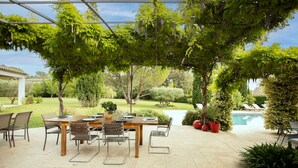 Un patio al aire libre con una mesa y sillas de madera, rodeado de exuberante follaje y una piscina.