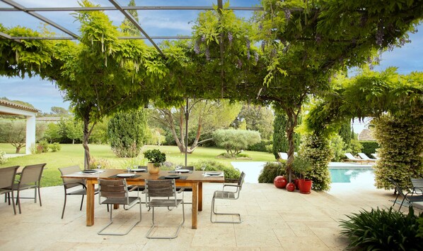 An outdoor patio with a wooden table and chairs, surrounded by lush greenery and a pool.