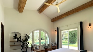 Un dormitorio con una cama grande, un ventilador de techo, vigas de madera, un ventanal con vistas a la vegetación y una silla con cojín.