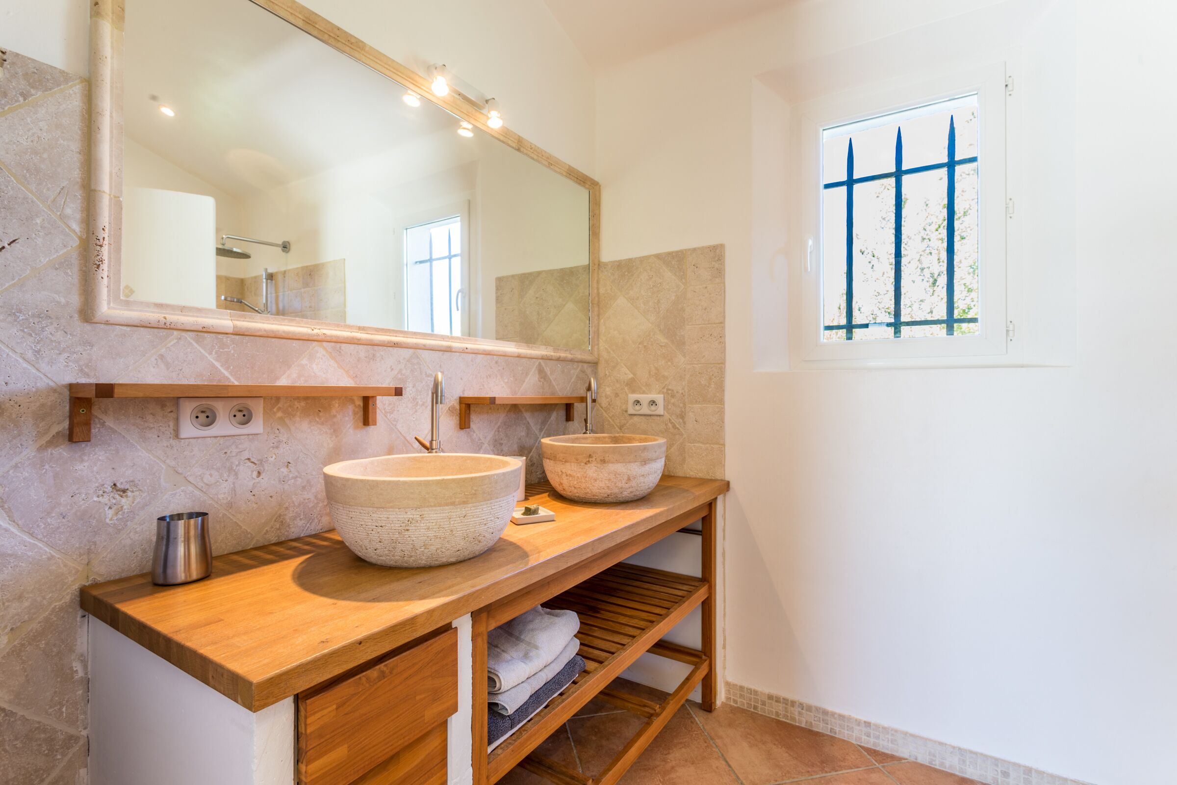 A bathroom with two large stone sinks, a wooden vanity, a mirror, and a window with blue bars.