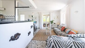 Een lichte, ruime woonkamer met een bank, een salontafel, en een keukenhoek met een aanrecht en kastjes.