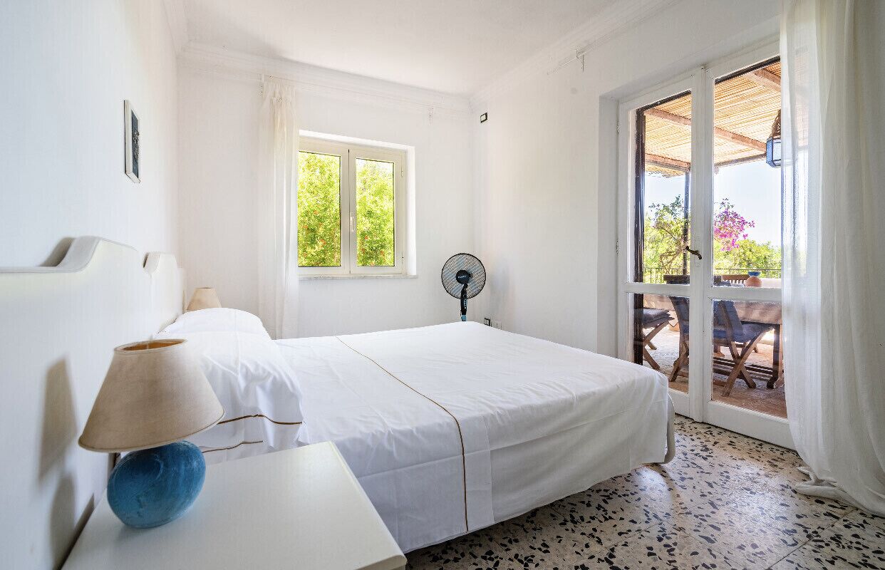 A bedroom with a bed, a bedside table with a lamp, a window, and a fan.