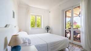 Een slaapkamer met een bed, nachtkastje met lamp, raam en ventilator.