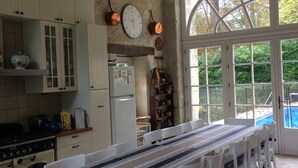 A rustic kitchen with a long dining table, white chairs, and a view of a swimming pool through large windows. - Large family house with pool in the heart of the Loire Valley (Langeais)