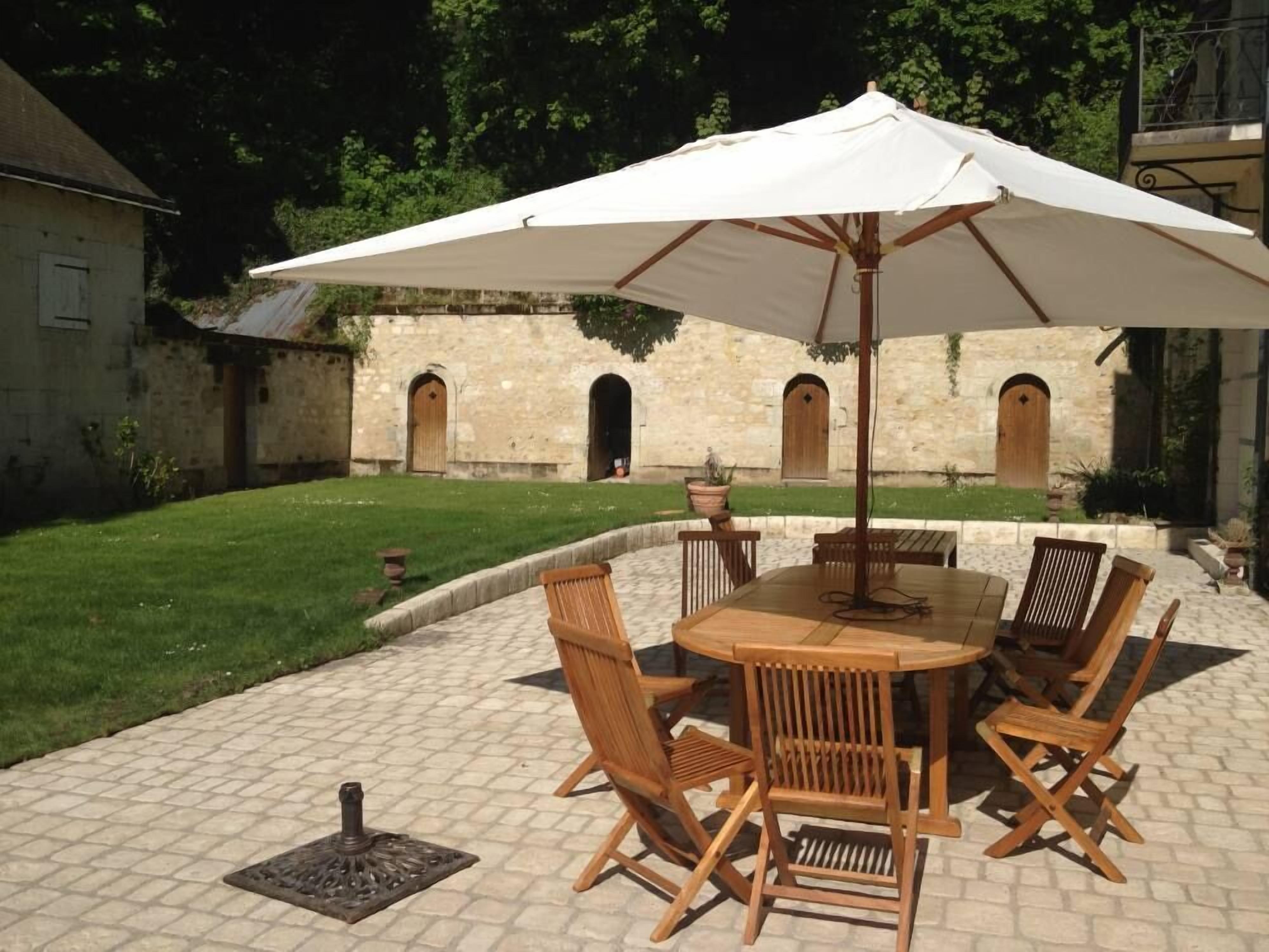 Une terrasse extérieure avec une table et des chaises en bois rondes, un grand parasol blanc, et un bâtiment en pierre avec des portes cintrées en arrière-plan.