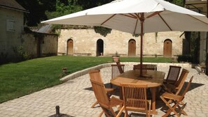 An outdoor patio with a round wooden table and chairs, a large white umbrella, and a stone building with arched doorways in the background. - Large family house with pool in the heart of the Loire Valley (Langeais)
