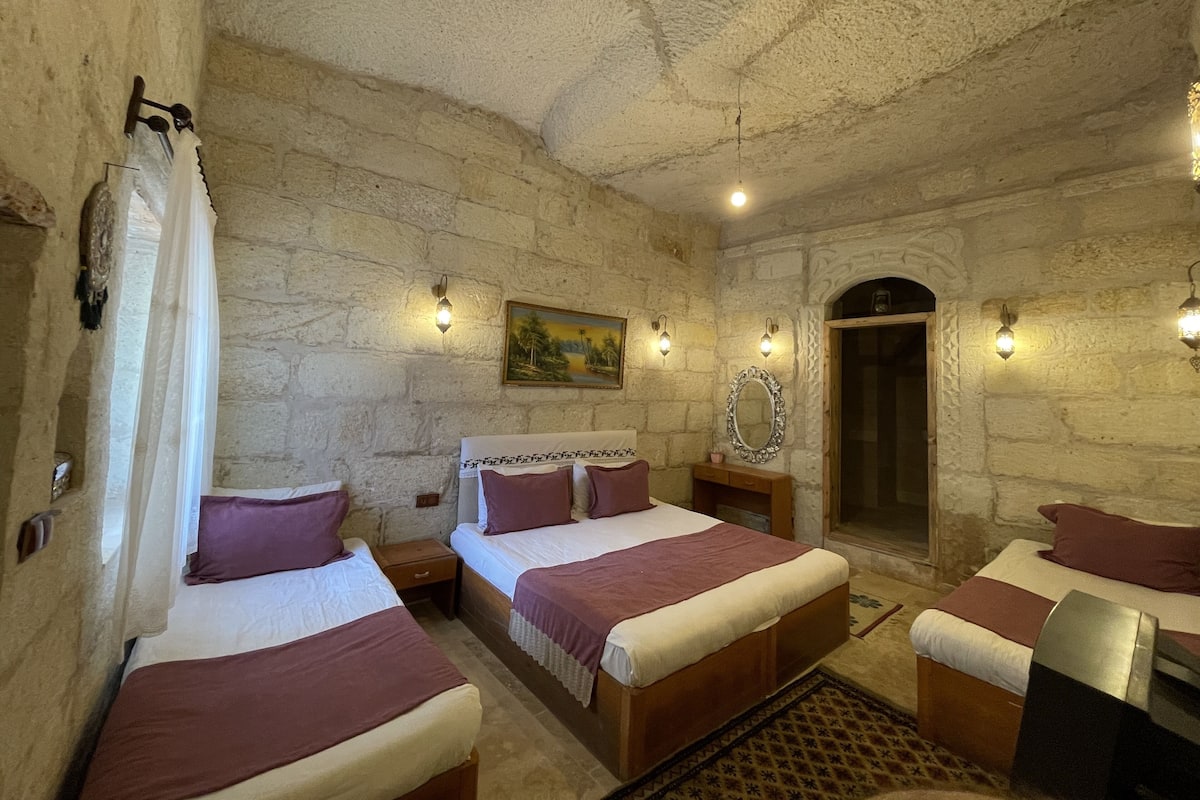 Habitación con tres camas, paredes de piedra y un cuadro. Se observan apliques de pared y una puerta.