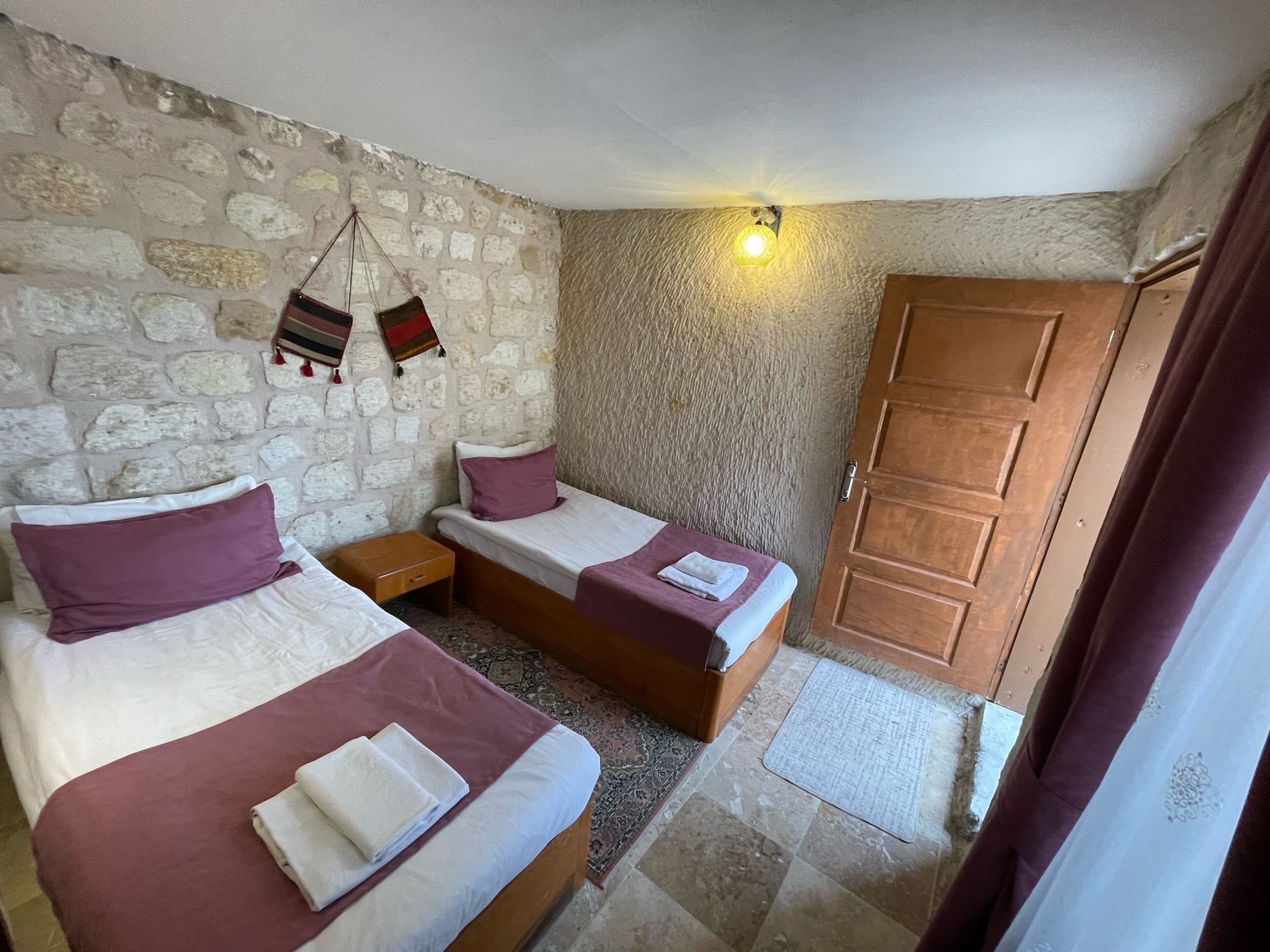 A room with two beds, a stone wall, a wooden door, and a small nightstand.