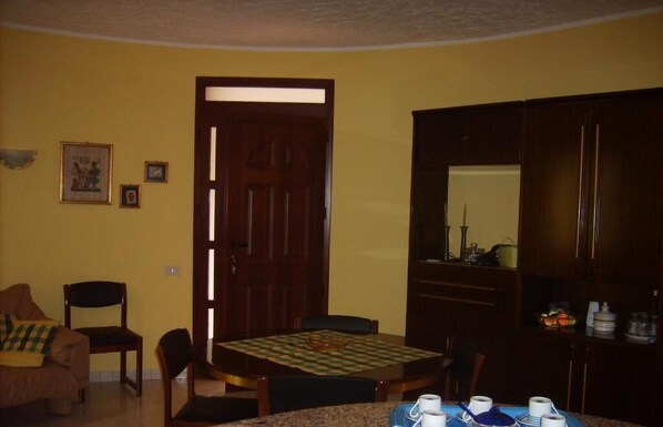 A dining area with a wooden table, chairs, and a checkered tablecloth. There is a door leading to another room, a small framed picture on the wall, and a cabinet with kitchen items.