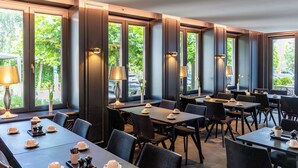 A restaurant with multiple tables and chairs, large windows with views of greenery, and table lamps. - &REPEAT Köln Airport (Cologne)
