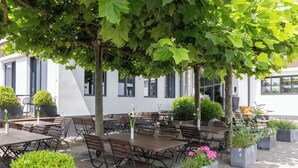 An outdoor patio with tables and chairs, surrounded by potted plants and trees. - &REPEAT Köln Airport (Cologne)