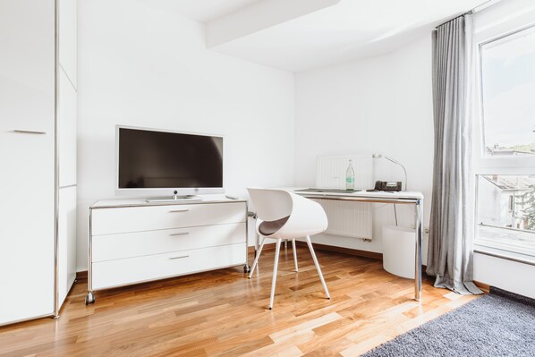 Una habitación moderna y minimalista con suelo de madera, muebles blancos, un televisor de pantalla plana, un escritorio con silla y una ventana con cortinas.