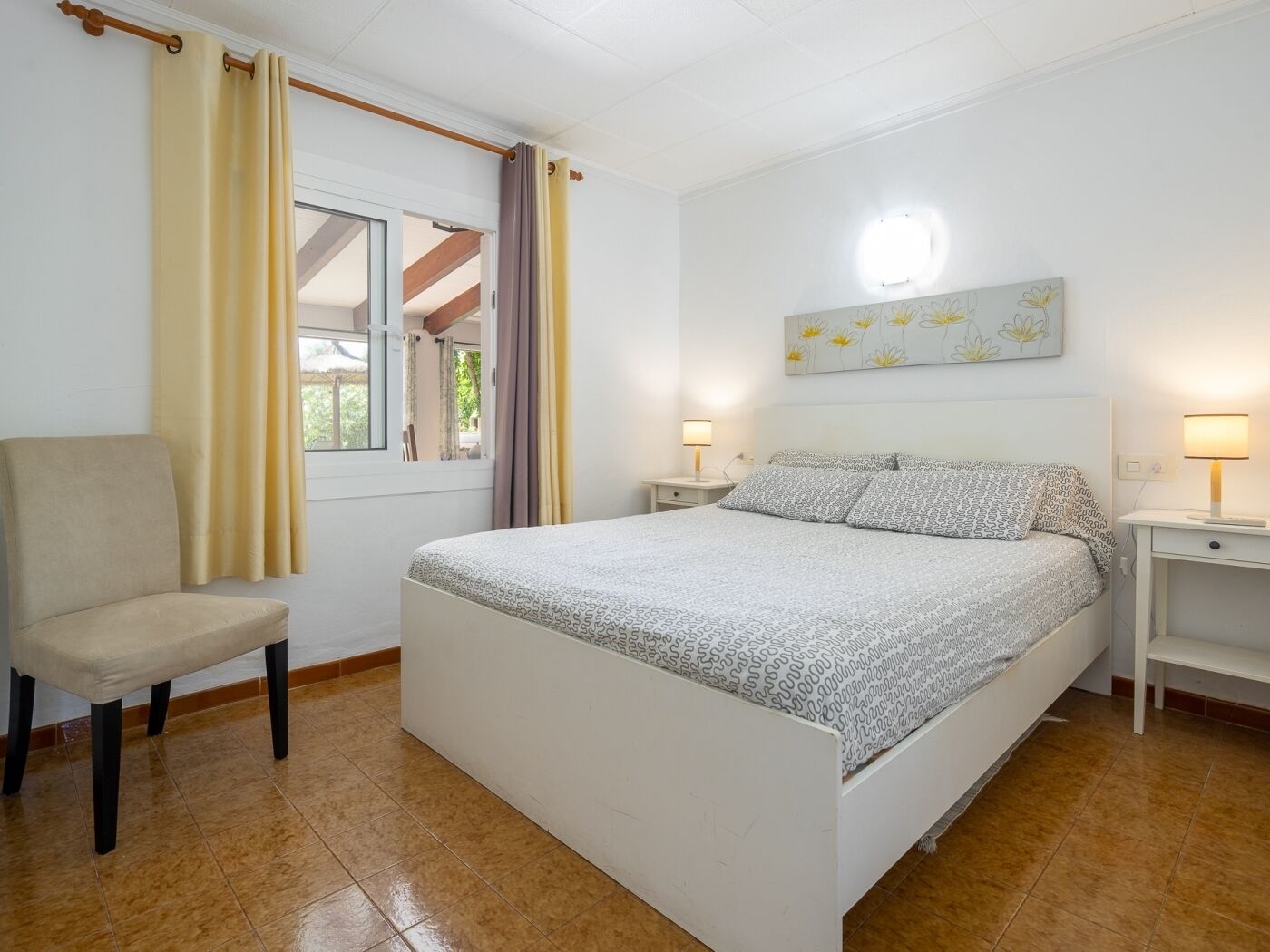 A bedroom with a bed, a chair, two bedside tables with lamps, a window with curtains, and a wall with a floral painting.