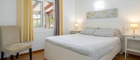 A bedroom with a bed, a chair, two bedside tables with lamps, a window with curtains, and a wall with a floral painting.