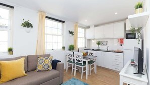 Eine moderne Wohnung mit Wohnzimmer, Essbereich und Küche. Sie verfügt über weiße Schränke, ein graues Sofa mit gelben Kissen und Topfpflanzen.