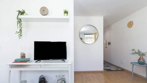 Ein modernes Wohnzimmer mit einem Flachbildfernseher auf einer weißen Konsole, einem runden Spiegel an der Wand und einem kleinen Tisch mit einer Pflanze.