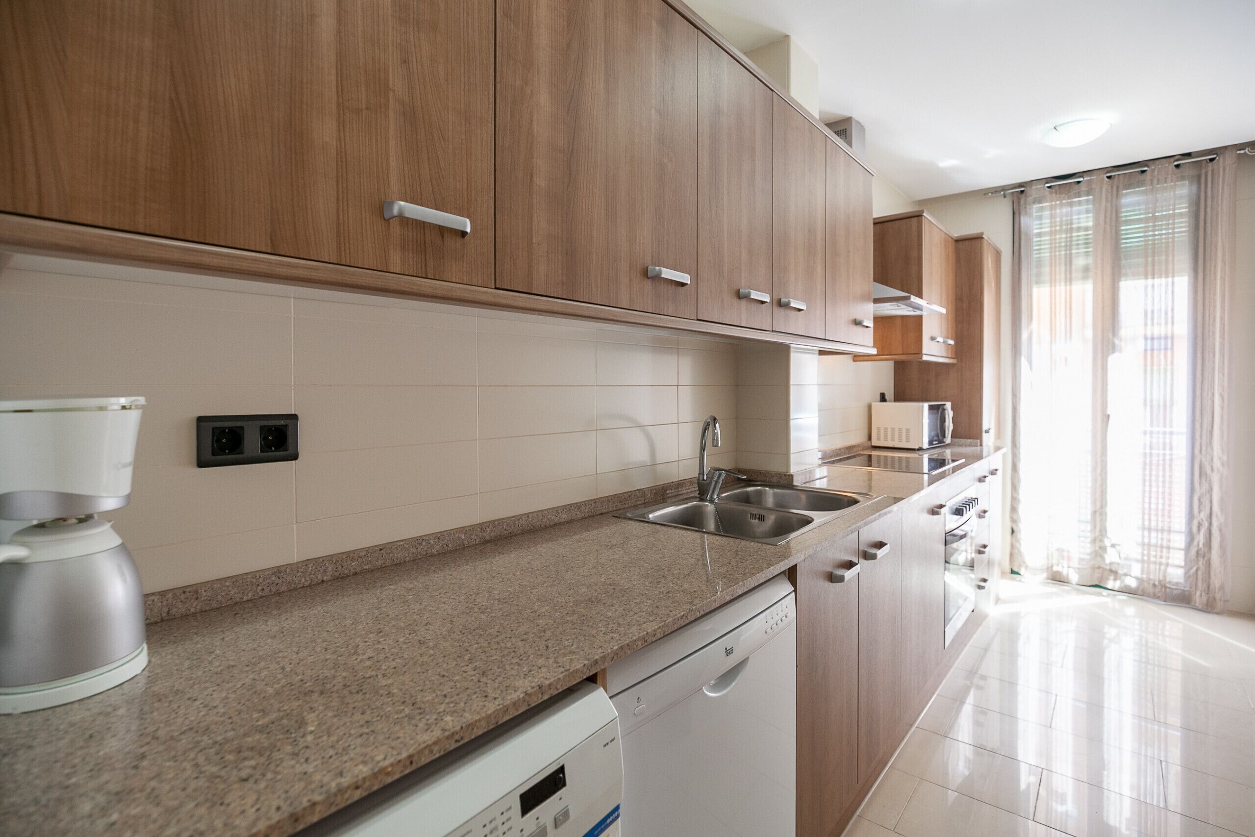 Een moderne keuken met houten kastjes, een granieten aanrechtblad, een ingebouwde vaatwasser en een raam met gordijnen.