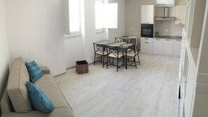 En moderne stue med sofa, spisebord med stole samt et køkken med skabe og hårde hvidevarer.
