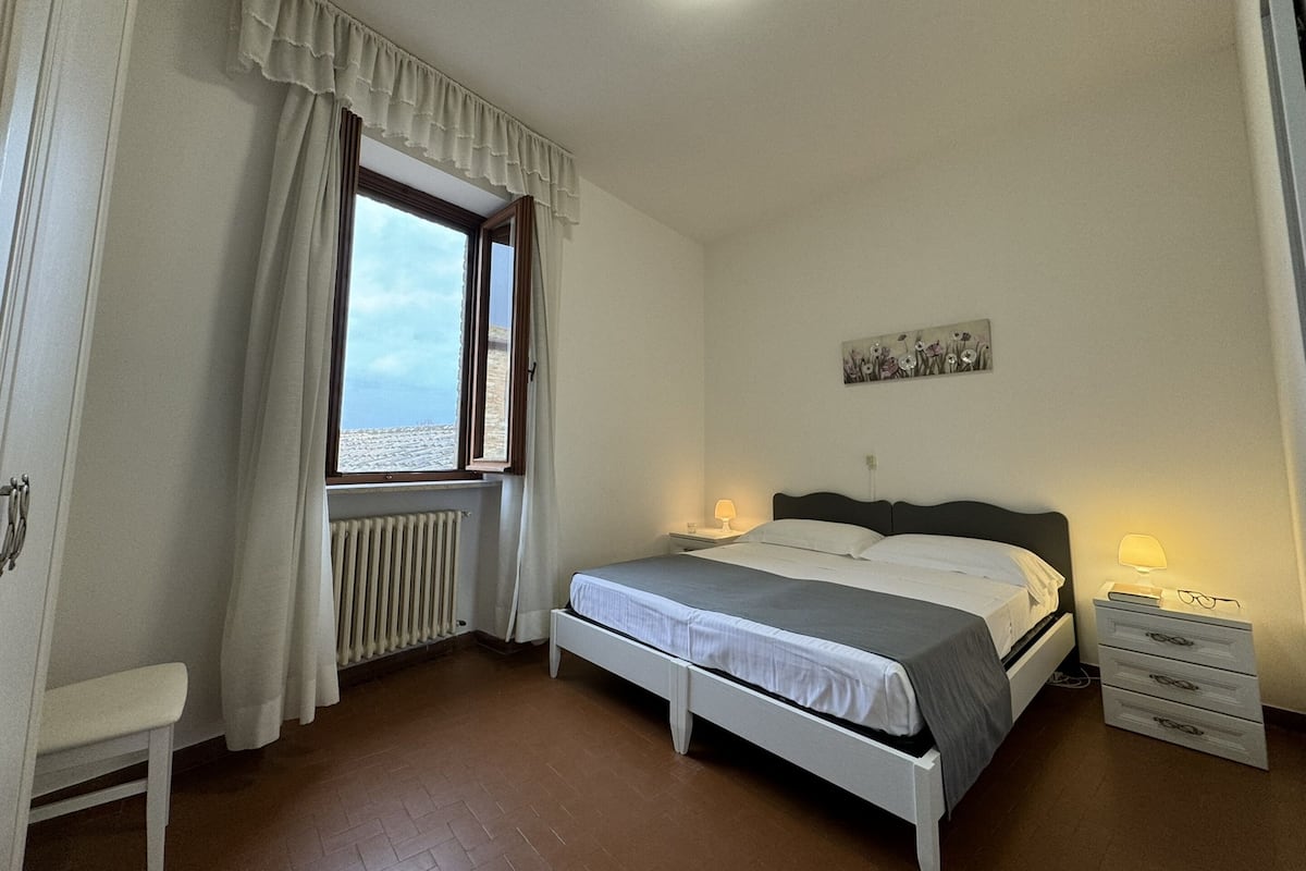 A bedroom with a bed, bedside tables, a radiator, a window with curtains, and a view of the sea.