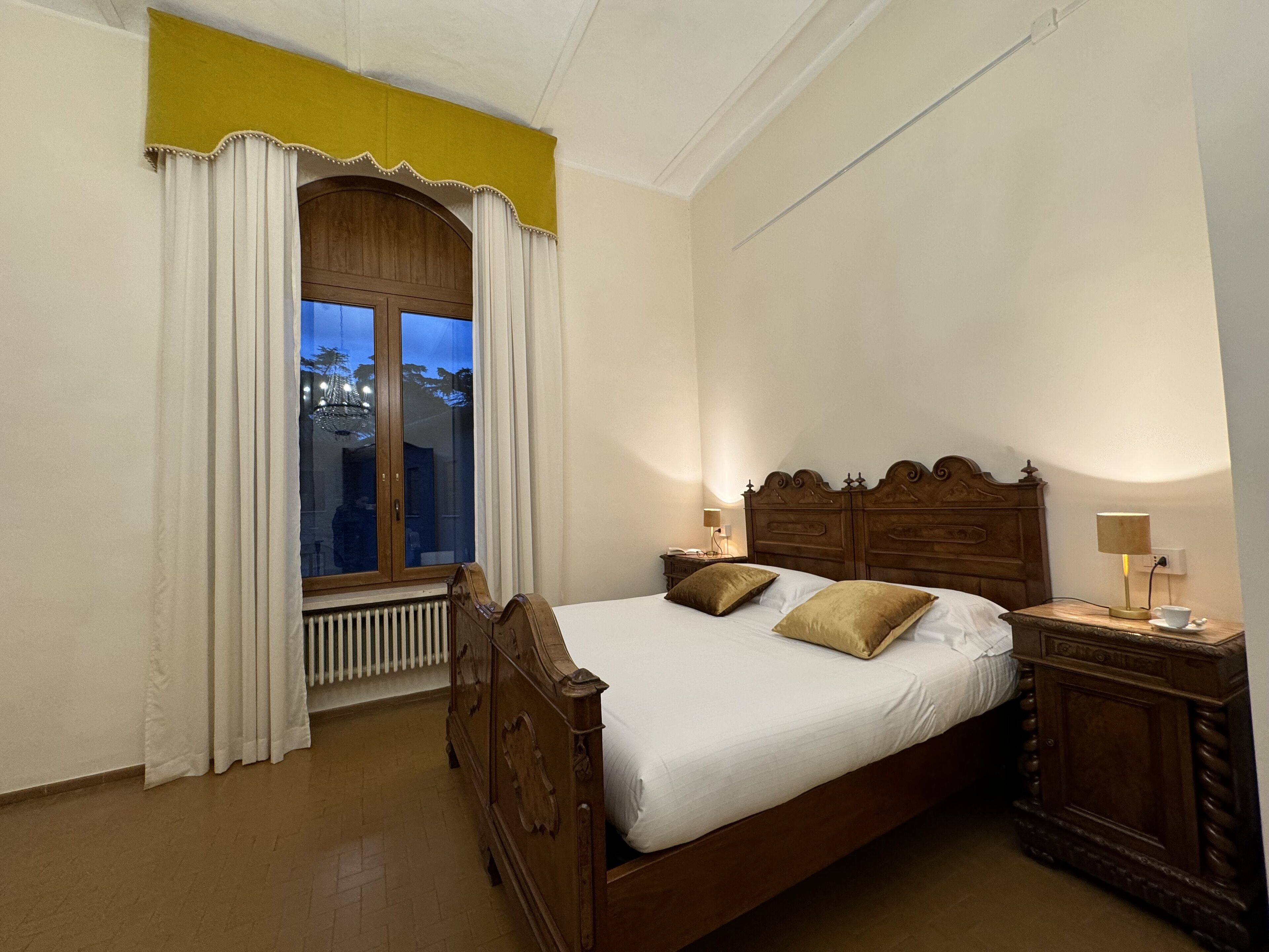 Un dormitorio con una cama de madera, dos mesitas de noche, una ventana con cortinas y un radiador.