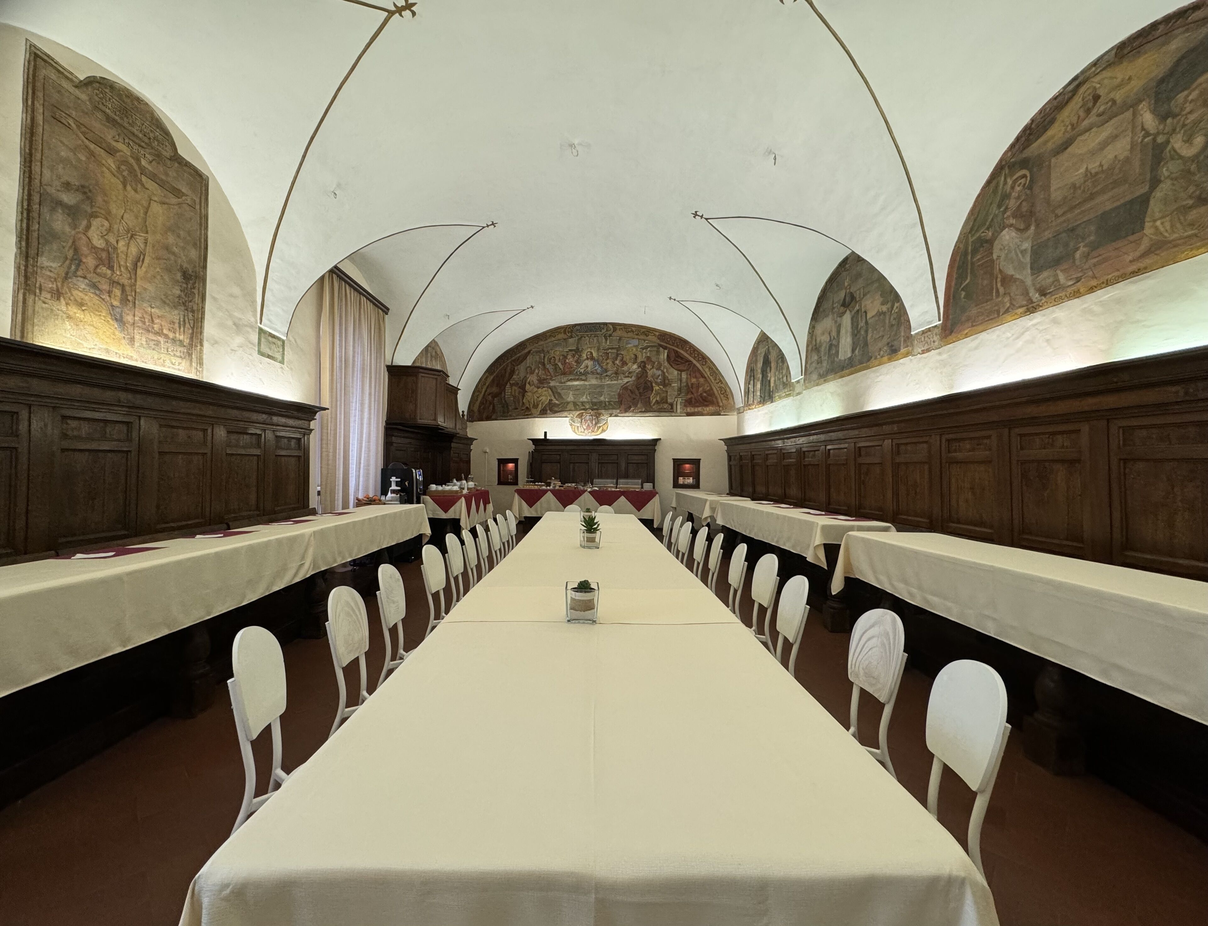 Een lange tafel met witte stoelen in een ruimte met gewelfde plafonds en fresco's op de muren.