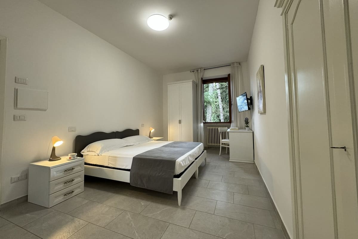 A bedroom with a bed, bedside tables, a desk, a chair, a TV, and a window with curtains.