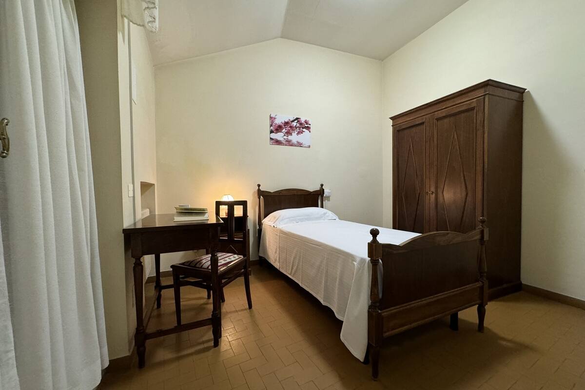 A bedroom with a single bed, a wooden wardrobe, a desk with a chair, and a wall-mounted lamp.