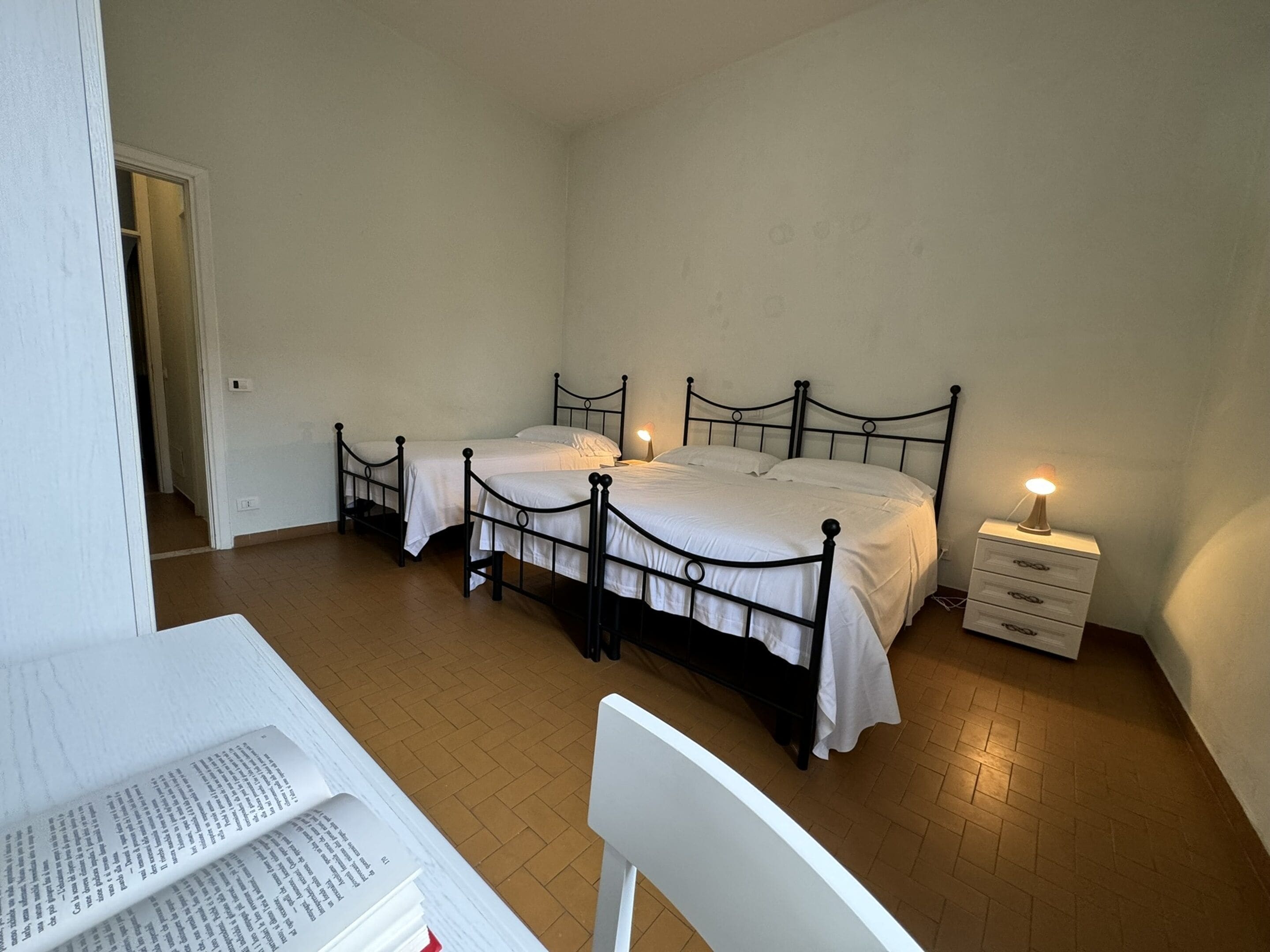 Una stanza con due letti singoli, una scrivania bianca con una sedia e un comodino con una lampada.