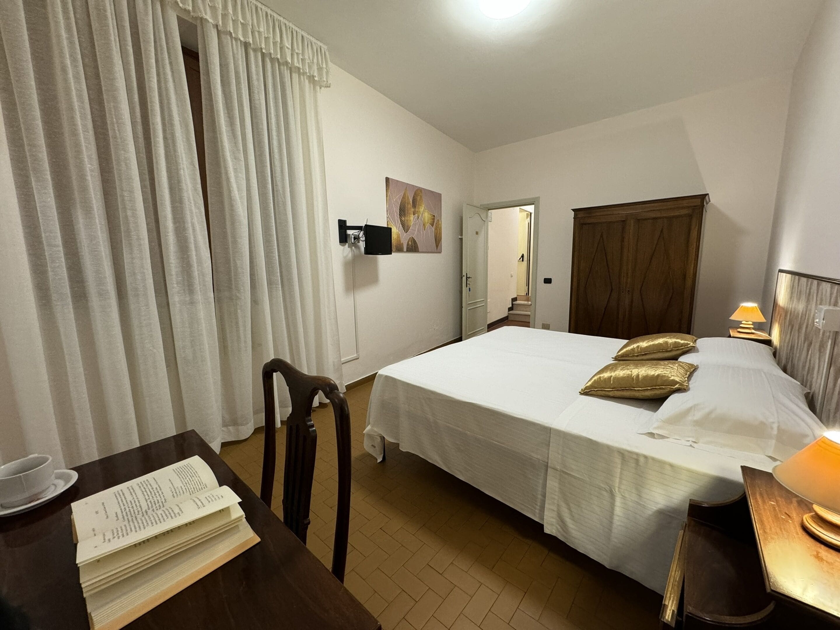 A hotel room with a bed, a desk with a book, a chair, a bedside table with a lamp, and a door leading to another room.