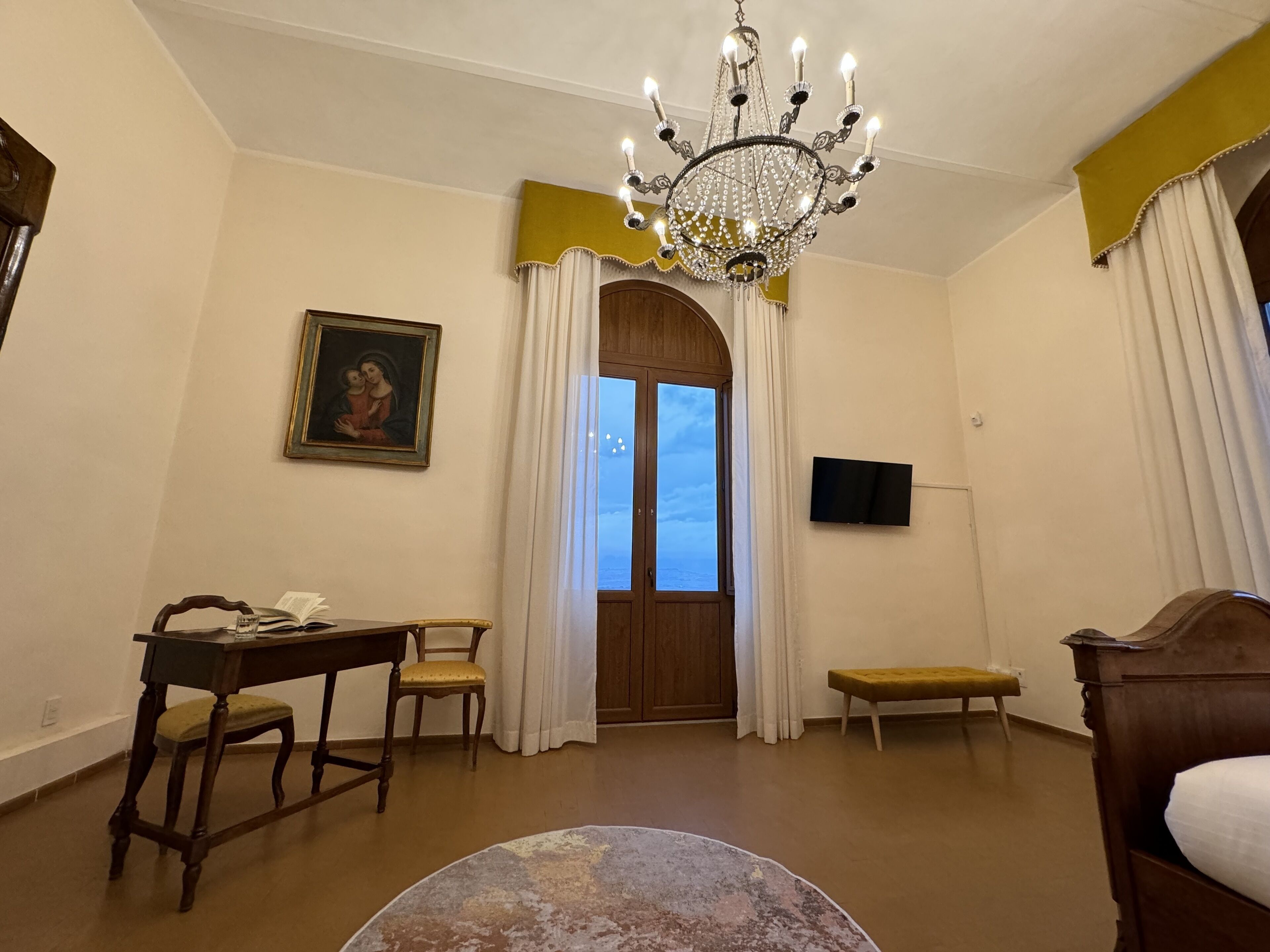 A room with a wooden desk, a chair, a bench, a television, and a chandelier.