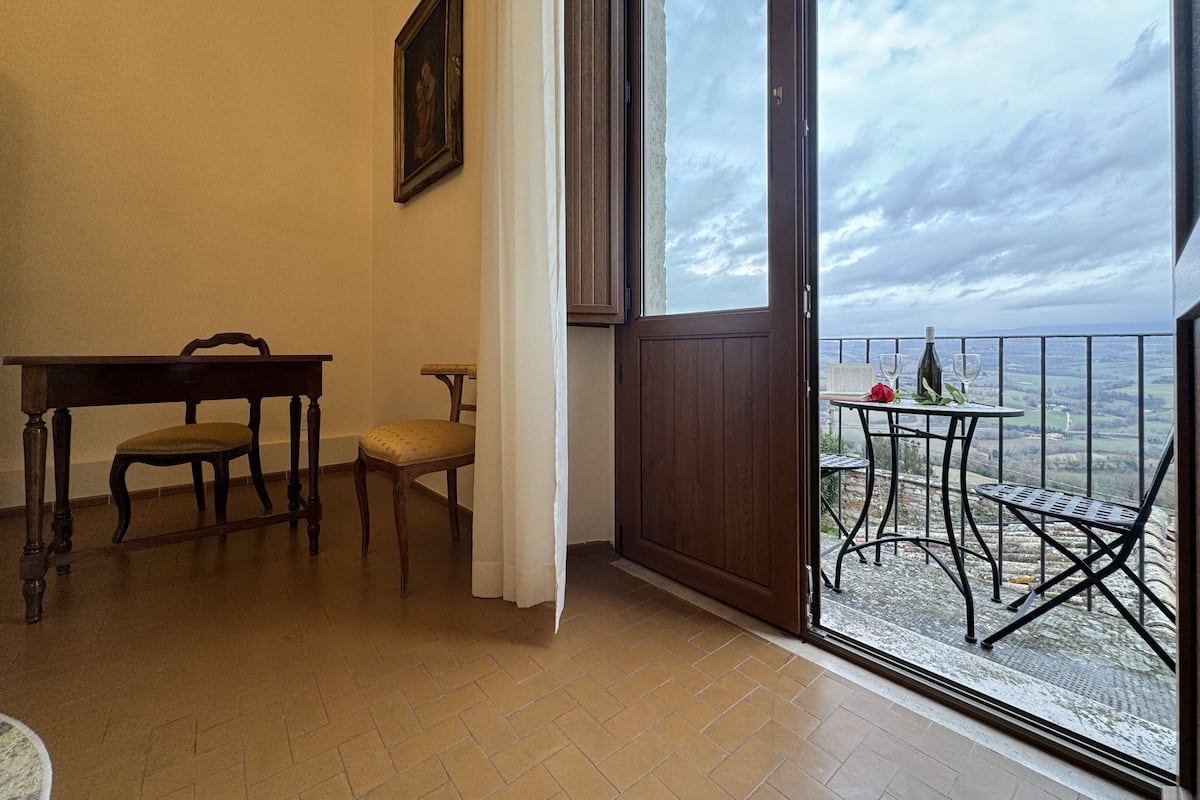 A room with a wooden table and two chairs, a door leading to a balcony with a table and chairs, and a framed picture on the wall.