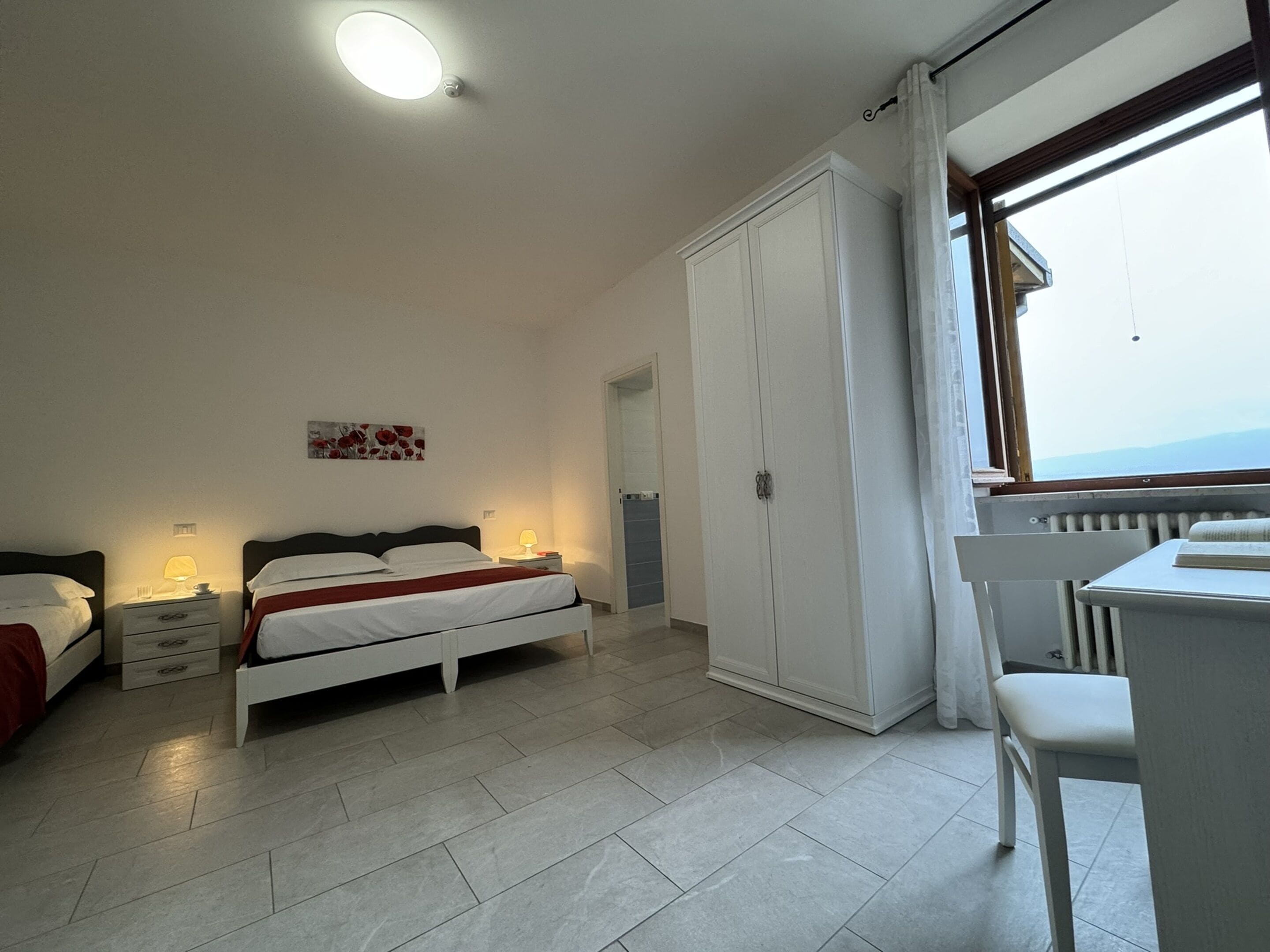A bedroom with a bed, bedside tables, a wardrobe, a desk, and a window with a view.